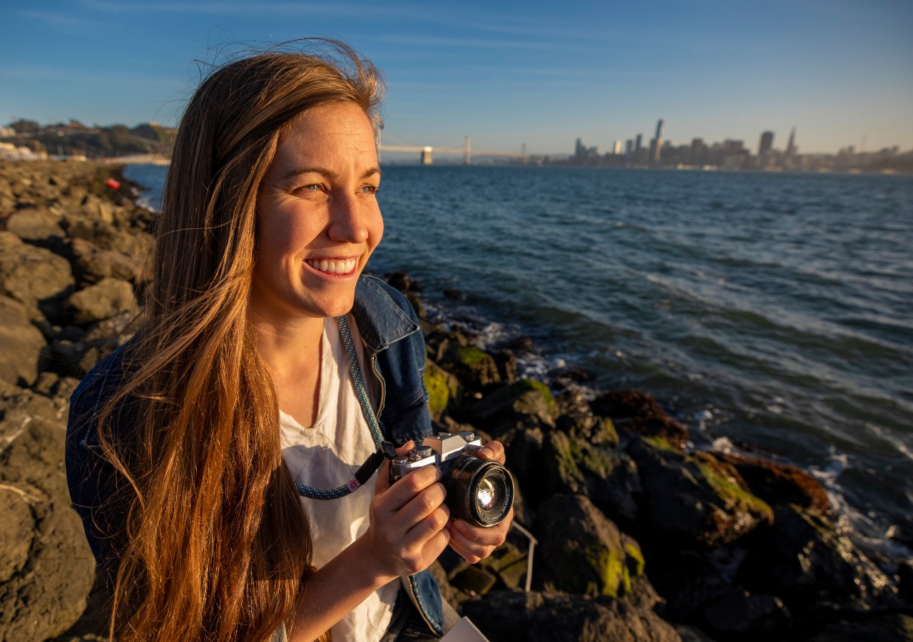Hope poses with a camera in front of Oakland Bridge
